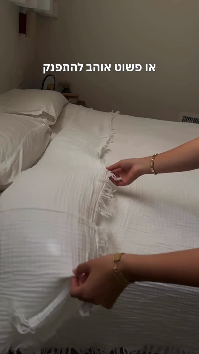 Hands straightening white gauze throw with fringe on neatly made linen bed in cozy minimalist bedroom.