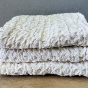 Cream waffle-knit cotton towels folded in a stack on a wooden surface.