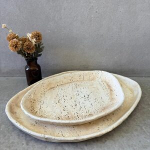 Handmade rustic stoneware plates with speckled texture, stacked on concrete, with dried flowers in amber glass vase.