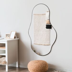 Asymmetrical wall mirror with black frame in minimalist Scandi room, beside white desk and woven pouf on wooden floor.