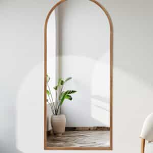 Full-length arched wall mirror with wooden frame reflecting potted plant in bright modern minimalist interior.