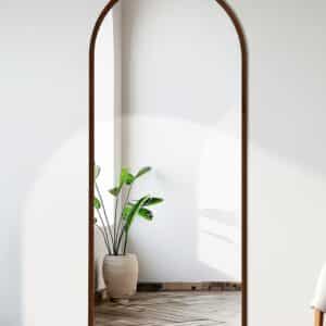 Full-length arched wall mirror with wood frame reflecting potted plant in bright minimalist room, herringbone floor.