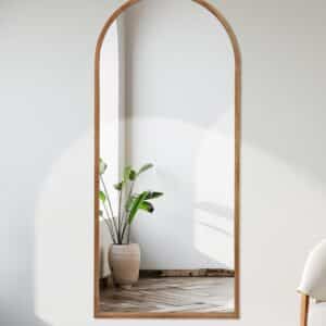 Arched full-length mirror with wood frame in minimalist room, reflecting potted plant and natural light, modern home decor.