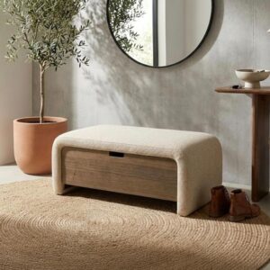 Beige upholstered storage bench in modern entryway with round mirror, jute rug, olive tree and terracotta planter.