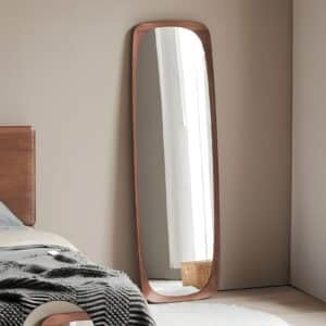 Full-length mirror with walnut frame leaning in modern neutral bedroom near window and bed with gray throw