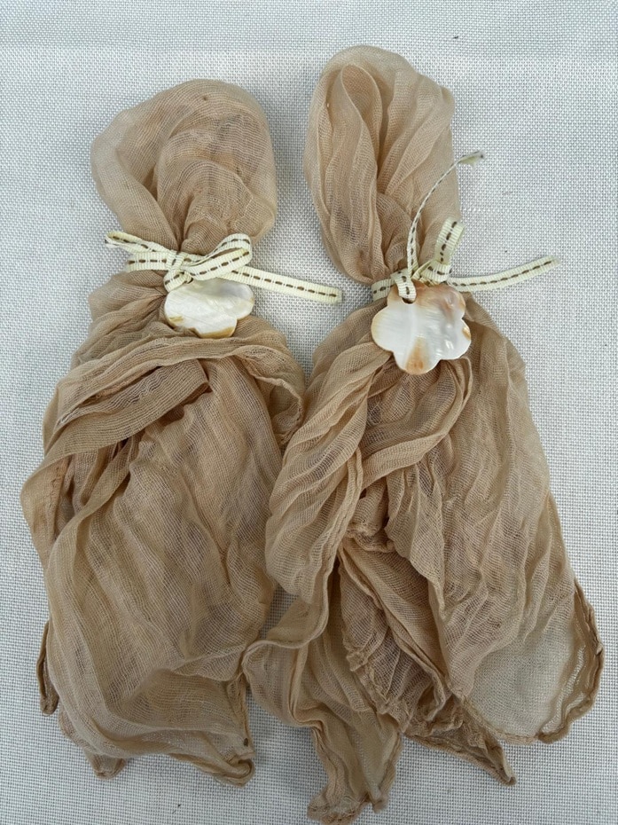 מפית בד וחבק (1) Pair of beige gauze napkins tied with ribbon and mother-of-pearl shell charms on a neutral linen background