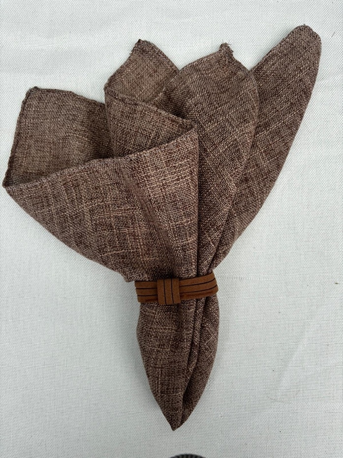 מפית בד וחבק (5) Rustic brown linen napkin folded with leather ring on neutral tablecloth, farmhouse table setting decor.