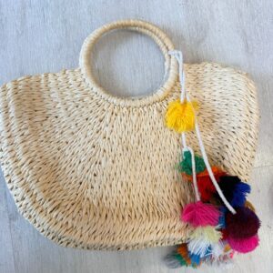 Woven straw tote handbag with round handles and colorful pom-pom tassels, boho summer beach bag on light wood floor